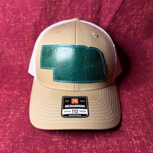 Puede incluir: Gorra de camionero beige y blanca con un parche de cuero verde con la forma de Nebraska. La gorra tiene una visera curva y un cierre de broche ajustable. La gorra Richardson 112 es un estilo clásico.