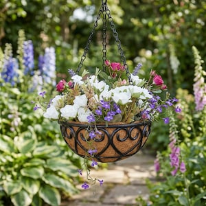 Può includere: Un cestino sospeso pieno di fiori bianchi, rosa e viola. Il cestino ha una cornice decorativa in metallo nero ed è sospeso da una catena nera. Lo sfondo presenta un giardino con varie piante e un sentiero di pietra.