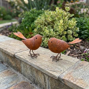 May include: Two rusty-brown metal bird sculptures with orange tail feathers and thin wire legs stand on a stone ledge. The birds face each other, set against a backdrop of green and yellow foliage, perfect for garden decor.