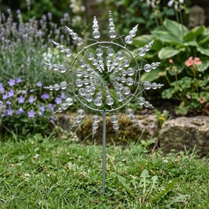 May include: A decorative garden spinner with a circular design. The spinner features clear glass beads and a silver metal frame. It is set in a grassy area with flowers and rocks in the background. The spinner is designed to catch the wind and create a visual display.