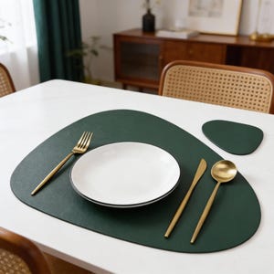 Op de afbeelding: Eettafelopstelling met een donkergroene placemat, een witte bord met een zwarte rand en goudkleurig bestek. Een bijpassende onderzetter en een gouden vork, mes en lepel maken de elegante opstelling compleet.