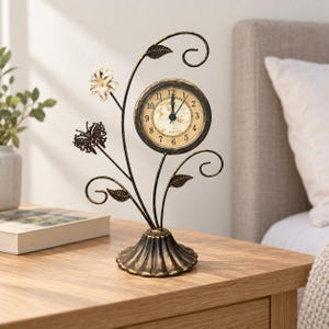May include: A black metal table clock with a distressed gold finish, featuring a round clock face with black numerals and hands. The clock is supported by a decorative stand shaped like a vine with metal leaves, a small flower, and a butterfly.