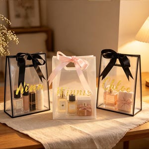 Puede incluir: Tres bolsas de regalo transparentes con lazos negros y rosas, que contienen productos de belleza. Las bolsas tienen nombres con purpurina dorada: "Winnie" y "Chloe". Se exhiben sobre una mesa de madera con un camino de mesa de color neutro.