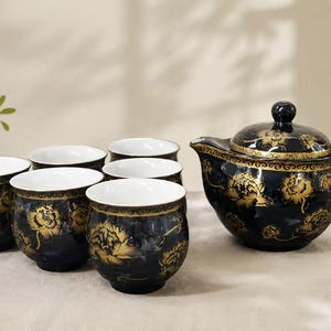 May include: A tea set consisting of a teapot with a lid and five matching cups. Each piece is made of black ceramic with a glossy finish, featuring intricate gold floral patterns and a white interior. The teapot has a curved handle and spout.