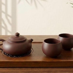 Peut inclure: Une théière en argile brun foncé avec un couvercle assorti et deux tasses à thé arrondies disposées sur un plateau de service rectangulaire en bois. L'ensemble présente une finition mate lisse et un petit vase en céramique claire.