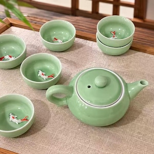 May include: A light green ceramic tea set consisting of a round teapot with a lid and five small tea cups. Each cup features a small, three-dimensional white and orange koi fish figurine attached to the interior base of the smooth, glazed vessel.