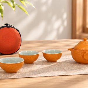 May include: A tea set consisting of three small bowls and a lidded teapot, all featuring a textured orange exterior and a smooth light blue interior. A round, orange zippered carrying case with a black trim is positioned behind the tea bowls.