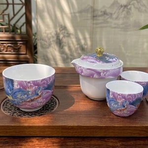 Puede incluir: Un juego de té de cerámica que incluye un gaiwan con un pomo dorado y tres tazas a juego. Cada pieza está decorada con un vibrante motivo de dragón en tonos morados y azules sobre un fondo blanco, dispuesto sobre una bandeja de madera oscura.