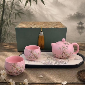 May include: A pink ceramic tea set consisting of a teapot and two matching cups, all decorated with delicate white and brown floral branch patterns. The set is arranged on a rectangular tray with a floral design, placed before a green storage box.