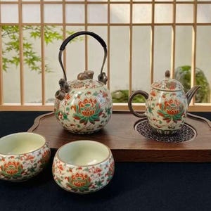 Peut inclure: Un service à thé en céramique comprenant une théière ronde avec une anse pivotante en métal foncé, une théière plus petite avec couvercle et deux bols assortis. Chaque pièce est ornée d'un motif floral coloré de lotus rouges et verts sur fond crème.