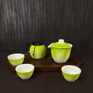 May include: A ceramic tea set featuring a lidded teapot, a small lidded jar with a dark brown lid and cord, and three small tea cups. Each piece has a textured, light green exterior resembling the leaves of a cabbage with white interiors.