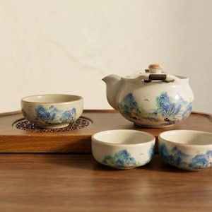 May include: A ceramic tea set consisting of a teapot with a lid and three small tea cups, all featuring a hand-painted blue and green mountain landscape design on a cream-coloured base. The set is arranged on a wooden serving tray.