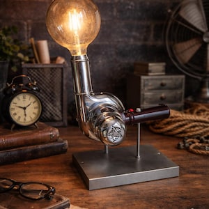 Op de afbeelding: Een unieke bureaulamp in steampunk-stijl met een grote Edison-lamp. De lamp is gemaakt van gepolijste metalen onderdelen, waaronder een turbine-achtige behuizing en een vierkante basis. Het warme licht verlicht het houten bureau.