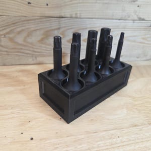 May include: A black tool holder with multiple black tools inserted. The tools are of varying heights and shapes, all with a similar cylindrical base. The holder is rectangular and sits on a wooden surface.