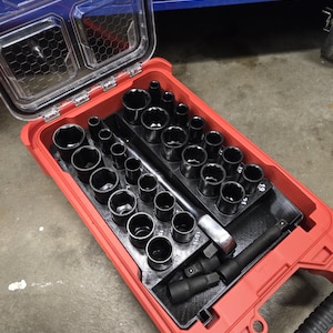 May include: A red toolbox with a clear lid, filled with a set of black sockets and a ratchet. The sockets are arranged in rows and sizes. The toolbox is open, revealing the tools inside. The brand name "PACKOUT" is visible on the lid.