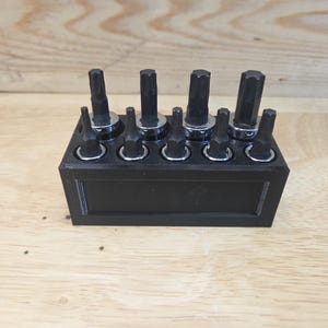 May include: A black tool organizer holding various hex bit sockets. The sockets are arranged in a row, with a mix of sizes and shapes. The organizer is rectangular and sits on a wooden surface.