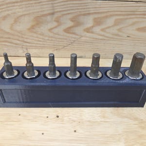 May include: A dark blue 3D-printed tool holder with eight metal hex bits. The bits are arranged in a row, each set in a circular recess. The holder sits on a light brown wooden surface.