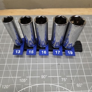 Puede incluir: Cinco vasos de metal de color plateado dispuestos en fila sobre un soporte organizador de plástico negro y azul. Las pestañas azules están marcadas con los números 13, 15, 16, 17 y 19, indicando el tamaño específico de cada herramienta.