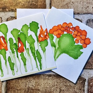 May include: Two square greeting cards with watercolour floral designs. One card features orange flowers and green leaves, while the other showcases orange flowers and green stems. The cards are on a white background.