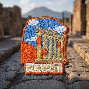 May include: An embroidered patch featuring a blue sky with white clouds, a tan and orange ancient temple with columns, and the text POMPEII in tan letters on an orange background, all outlined with a thick orange border.