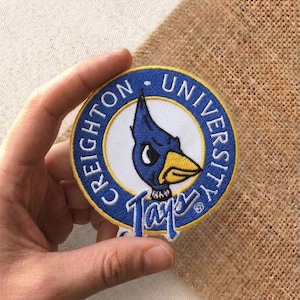May include: Embroidered patch featuring the Creighton University logo. The patch is circular with a blue and yellow border, and the text "CREIGHTON UNIVERSITY" is embroidered around the edge. The center features a blue bird with a yellow beak. The word "Jays" is embroidered below the bird.