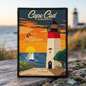 May include: A rectangular embroidered patch featuring a lighthouse with a red top and white base, a sailboat on blue water, and a sunset sky. The text Cape Cod Massachusetts is stitched in gold at the top. A green tree and bicycle are visible.