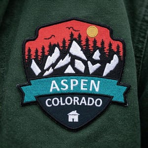May include: A shield-shaped embroidered patch on a dark green fabric background. The design features a red sky with a yellow sun, black pine trees, and white mountain peaks. A teal banner displays the white text ASPEN COLORADO above a small house icon.