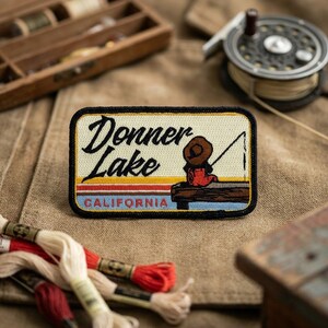 May include: Rectangular patch with the words "Donner Lake CALIFORNIA" in black script. The patch features a cartoon image of a person fishing off a dock, with a brown hat and red clothing. The background has horizontal stripes of yellow, orange, red, and blue.