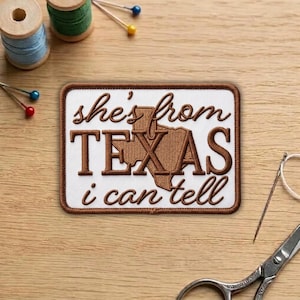 May include: A rectangular patch with the text "she's from TEXAS i can tell" in brown embroidery. The patch features a brown outline of the state of Texas. The patch is on a white background with a brown border.