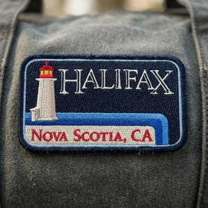 May include: A rectangular embroidered patch featuring a white lighthouse with a red top on the left side. The text HALIFAX is in white, and the text NOVA SCOTIA, CA is in red on a light background, all set against a dark blue background.