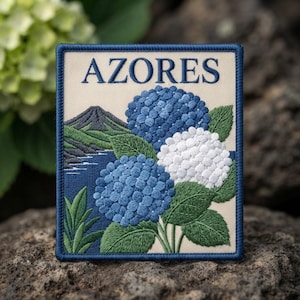 May include: A rectangular embroidered patch with a cream background and a dark blue border. The text AZORES is stitched in dark blue at the top. The design features a dark green mountain, a blue sea, and clusters of blue and white hydrangea flowers.