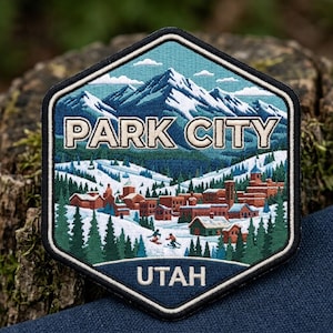 May include: A hexagonal embroidered patch featuring a snowy mountain landscape with the text PARK CITY in white letters at the top and UTAH in white letters at the bottom. The scene includes brown buildings, green pine trees, and two skiers.