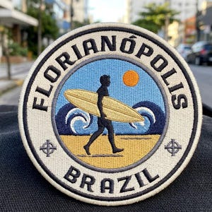 May include: A circular embroidered patch featuring a black silhouette of a person carrying a yellow surfboard on a beach. The design includes blue ocean waves, an orange sun, and the text FLORIANÓPOLIS and BRAZIL in black lettering.