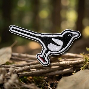 May include: An embroidered patch shaped like a magpie bird, featuring a black body with white wing and tail details, a white outline, and a small red accent on the foot. The design is rendered in a flat, graphic style with visible stitching.