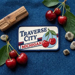 May include: A rectangular embroidered patch with a cream background and dark blue border. It features the text "TRAVERSE CITY" in blue and "MICHIGAN" in red, accompanied by an embroidered graphic of two red cherries on a light blue circle.