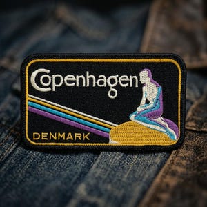 May include: Embroidered patch featuring the word "Copenhagen" in white, above a mermaid illustration. The patch has a black background with a yellow border and the word "DENMARK" at the bottom. The mermaid is in shades of blue and purple.
