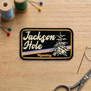 May include: A black embroidered patch with the words "Jackson Hole Wyoming" in a cream-colored font. The patch features a rainbow design and a detailed tree illustration. Sewing supplies, including thread spools and needles, are visible.