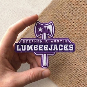 May include: A purple and white embroidered patch featuring the text "STEPHEN F. AUSTIN LUMBERJACKS." The patch has a stylized axe design at the top with a star and tree graphic. The patch is held in a hand.