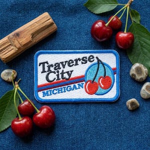 May include: A rectangular embroidered patch with a white background and a blue border. The text reads Traverse City MICHIGAN in black and blue. A graphic of two red cherries on a light blue circle is featured on the right side of the patch.