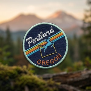 May include: A circular embroidered patch with a dark blue background. It features the white text Portland, the orange text OREGON, a light blue leaping deer silhouette, and a diagonal stripe pattern in orange, yellow, and light blue.