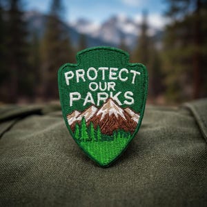 May include: A shield-shaped embroidered patch featuring a green background with the white text PROTECT OUR PARKS above a graphic of brown and white snow-capped mountains and a forest of green pine trees at the base of the mountain range.