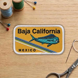 May include: Embroidered patch with the text "Baja California" and "MEXICO" on a yellow background. A teal fish is featured in the center, with blue and white stripes. The patch is rectangular with rounded corners.