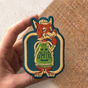 May include: Embroidered patch featuring a cartoon fox wearing a hat and a green shirt with the word "OHIO". The patch has a teal background with a gold border and is likely a souvenir or collectible item.