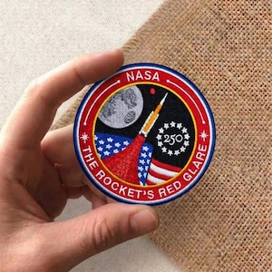 May include: Embroidered patch featuring a rocket launch with a red and white trail against a black background with a moon and stars. The patch includes the text "NASA" and "THE ROCKET'S RED GLARE". The patch is round with a blue and red border.