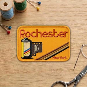 May include: Embroidered patch with the word "Rochester" in orange script. The patch features a film roll graphic and the text "New York" on a yellow background. The patch has a retro design with brown and black stripes.