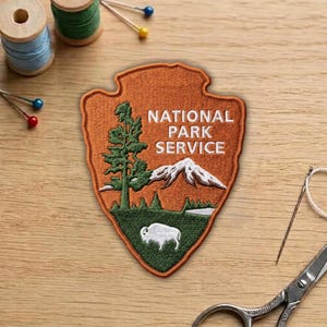 May include: Embroidered patch in the shape of an arrowhead, featuring the text "NATIONAL PARK SERVICE." The patch has a detailed design of a tree, mountains, and a bison. The patch is orange, green, and white.