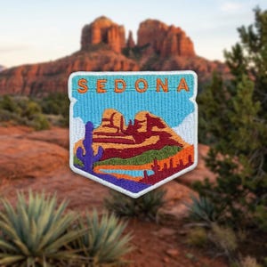 May include: A shield-shaped embroidered patch featuring a landscape of red rock formations under a light blue sky. The word SEDONA is stitched in orange at the top. The design includes a purple saguaro cactus, green desert vegetation, and orange ground.