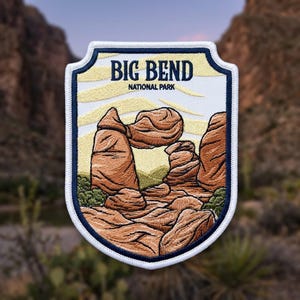 May include: A shield-shaped embroidered patch featuring a landscape of Big Bend National Park. The design displays brown rock formations, green desert vegetation, and a pale yellow sky. The text BIG BEND NATIONAL PARK is stitched in dark blue.