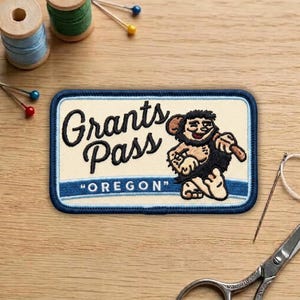 May include: A rectangular patch with a light beige background and a dark blue border. The patch features the words "Grants Pass" in a script font, and "OREGON" in quotation marks. A cartoon caveman is also featured.