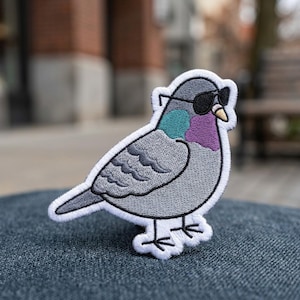 May include: An embroidered patch featuring a grey pigeon wearing black sunglasses. The bird has a teal and purple patch of color on its neck, a small yellow beak, and a thick white border outlining the entire shape of the bird against a dark background.